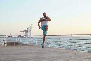 Photo of young masculine sporty bearded jumping guy doing morning exercises by the sea, warm-up before run, leads healthy active lifestyle. Fitness male model.