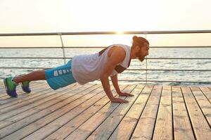 foto de joven barbado hombre haciendo Mañana ejercicios por el mar, haciendo Lagartijas, mantiene el tablón, calentar después correr, Guías sano activo estilo de vida. aptitud y sano concepto..