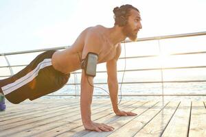 Portrait of young bearded sporty gay doing pushups, listening favorite songs on headphones, keeps the plank, doing morning exercises by the sea, warm-up after run, leads healthy active lifestyle. photo