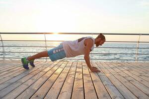 Photo of young sporty bearded man doing morning exercises by the sea, warm-up after run, doing pushups, keeps the plank, leads healthy active lifestyle. Fitness male model.