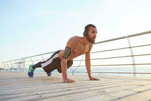 sporty bearded man listening favorite songs on headphones, doing morning exercises by the sea, warm-up after run, doing pushups, keeps the plank, leads healthy active lifestyle. Fitness male model. photo