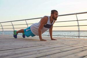 Young masculine sporty bearded guy doing morning exercises by the sea, warm-up after run, doing pushups, keeps the plank, leads healthy active lifestyle. Fitness male model. photo