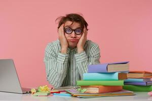 joven cansado hombre con gafas,sentado a un mesa con libros, trabajando a un computadora portátil, mira infeliz y triste, tristemente mira arriba y Sueños de dejando hogar como pronto como posible, aislado terminado rosado antecedentes. foto