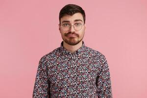 Close up of displeased young bearded man in glasses, wearing in colorful shirt, isolated over pink background and looking at camera with a raised eyebrow. People and emotions concept. photo