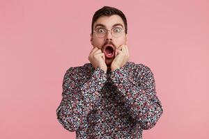 Portrait of young shocked bearded man in glasses, wearing in colorful shirt, with wide open mouth, bites finger nails. Isolated over pink background. People and emotions concept. photo