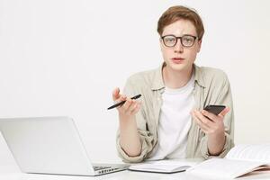 Guy student is sitting at the table, notebooks are lying in front of him and a laptop is standing, talks to someone, who is ahead of him, holds a phone and a pen in his hands photo