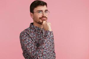 Portrait of young man in colorful shirt looking upwards, copy space on the right side, think about problem, while touches cheek, isolated over pink background. photo