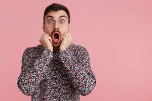 Studio shot of young screaming bearded man in glasses, wearing in colorful shirt, heard the shocking news, with wide open mouth. Isolated over pink background with copy space on the right side. photo