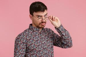 Close up of young man in colorful shirt, with confused face expression while, looking through glasses having doubts, isolated over pink background. photo