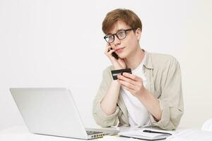 Studio shot of young man sits at a table and looks to the side through his glasses, holds a credit card in his hands, talks on the phone, a notebook pen and an open laptop are on the table photo