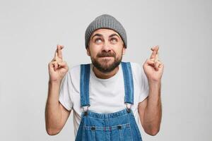 Prayers,wishing for good luck, hoping for forgiveness. Young humble man in a gray hat, white t-shirt, denim overall, keeps head and arms up and fingers crossed while standing against white background. photo