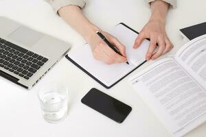 Close up of young man hands writing notes, making abstract, summary, opened a notebook on the table, there is a laptop near by, glass of water, phone with black screen, book and notebook photo