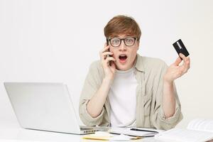 Young guy talking to a bank manager on the phone, looking at the camera through his glasses, holding a credit card, expression of bewilderment on his face, in confusion, panic, wide open mouth photo