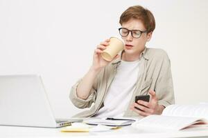 Young man is sitting at the table, drinking coffee, holding a phone in his hand, a notebook pen is on the table and laptop opened,looks on smart phone photo