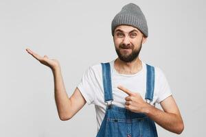 A man with a displeased squeamish unsatisfied facial expression looks into the camera, with his index finger shows towards to copy space on his palm, a white background photo