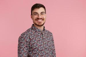 Close up of handsome cheerful bearded young man in glasses, wearing in colorful shirt, smiling and looking at camera, isolated over pink background with copy space on the right side. photo