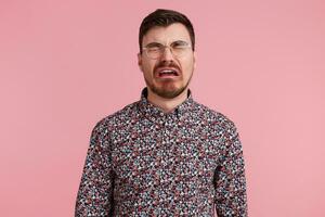 Portrait of a sad crying bearded man in glasses, wearing in colorful shirt, looks unhappy and upset. Isolated over pink background, people and emotions concept. photo