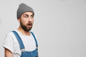 Close up of young man stands sideways with his eyes and mouth wide open, looks at the empty copy space. On his face an expression of surprise, wonder, amazement, isolated over white background photo