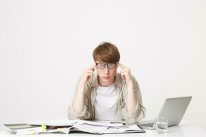 Young student sitting at the table, elbows put on the table, looks through his glasses with an indifferent or focused look, holds his fingers at the temples depicts a brainstorm or a suicide gesture photo