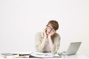 A young student guy is sitting at the table listening to someone on the phone, holding his head with his hand, looking away, boring uninteresting conversation, waiting for the operator to answer photo