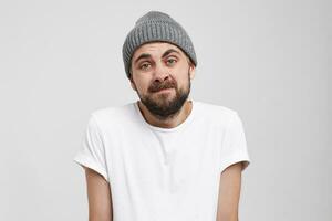 Confused young girl shrugging shoulders, having guilty look, feeling sorry for doing something wrong and making terrible mistake, dressed grey hat and white t-shirt, ,over white background photo