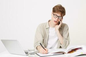 The student is engaged, sits at the table, talking on the phone, takes notes, looks into his notebook, consults with the group mate, takes notes photo