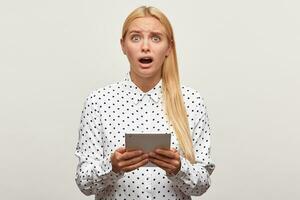 Shoot of a blonde young girl with tablet in hands, mouth open looks frustrated distressed upset because of some news or message wears white shirt with black dots, looking camera, on white background photo