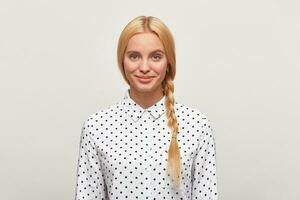 Beautiful diligent blonde woman with blue eyes, braid, feels happy, tenderly smiles, wears white shirt with black polka dots. Modest, excellent student, came to interview. Over white background photo