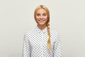 Disparo de un bonito rubia joven niña con natural maquillaje bueno piel pelo reunido en trenza extensamente sonrisas, usa blanco camisa con negro polca puntos, en un blanco antecedentes en el estudio. foto