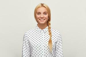 Portrait of a beautiful blonde young girl with natural make-up and well-groomed hair gathered in braid tenderly smiles, wears white shirt with black polka dots, on a white background in the studio. photo