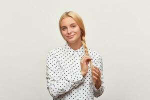 Beautiful diligent blonde woman with blue eyes, corrects keeps braid, feels happy, shows tenderness, wears white shirt with black polka dots. Over white background photo