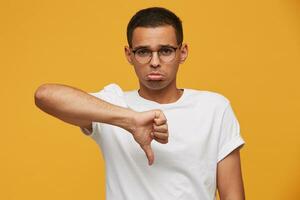 retrato de joven hombre en lentes mira triste, decepcionado, frustrado, disgustado con el resultado de el juego, dedo abajo, labios abadejo, usa blanco casual camiseta, en un amarillo antecedentes foto