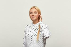 Portrait of beautiful diligent blonde woman with blue eyes, standing in a half-turn corrects hair braid, feels happy, shows tenderness, wears white shirt with black polka dots. Over white background photo