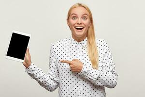 Blonde young girl looks happy delighted gladden amazed with tablet in hand, finger pointing on it, black screen looks camera for your message or advertisement,mouth open,on white background photo