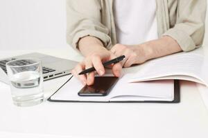 View from the top on young man with pen in hand making notes, opened a notebook on the table, laptop near by, glass of water, searching information on smart phone with black screen, book and notebook photo