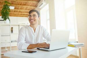 Indoor shot of smiling young businessman wears white shirt and glasses feels happy, using laptop and smartphone sitting at the table in office photo