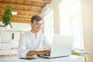 Indoor shot of happy attractive young businessman wears white shirt and glasses feels satisfied, using laptop and smartphone working at the table in office photo