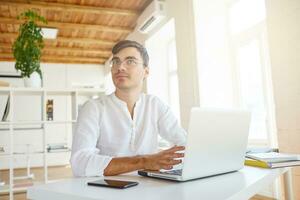 Closeup of handsome pensive young businessman wears white shirt and spectacles using laptop and smartphone sitting at the table and thinking in office photo