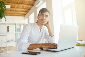 Indoor shot of pensive concentrated young businessman wears white shirt and spectacles using laptop and mobile phone thinking and working at the table in office photo