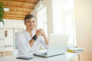 Portrait of happy successful young businessman wears white shirt and spectacles using laptop and cell phone sitting at the table in office and smiling photo