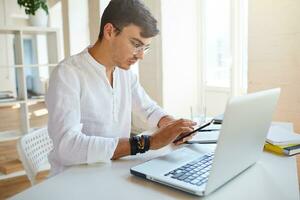 Portrait of concentrated handsome young businessman wears white shirt and spectacles using laptop and smartphone working at the table in office photo