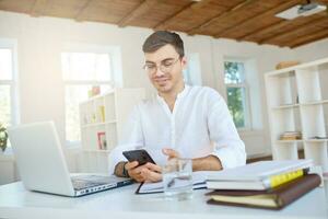 Closeup of smiling attractive young businessman wears white shirt and glasses with laptop and mobile phone feels happy and working at the table in office photo