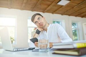 Portrait of thoughtful handsome young businessman wears white shirt and spectacles using laptop and smartphone working at the table in office photo