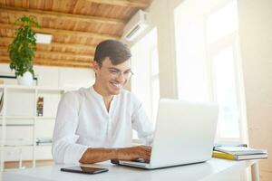 Portrait of cheeful confident young businessman wears white shirt and spectacles using laptop and smartphone working at the table in office and laughing photo