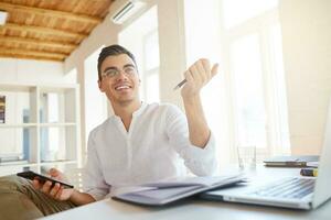 Closeup of smiling attractive young businessman wears white shirt and spectacles using smartphone and writing in notebook at the table in office photo