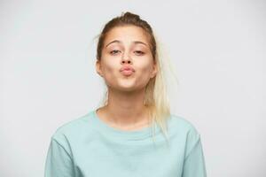 Closeup of playful attractive young woman with blonde hair and ponytail wears blue sweatshirt flirting and kissing isolated over white background photo