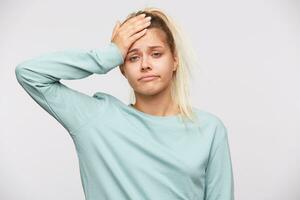 Portrait of upset depressed young woman with blonde hair and ponytail wears blue sweatshirt feels ill, touching her head and having headache isolated over white background photo