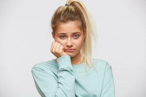 Portrait of unhappy disappointed young woman with blonde hair and ponytail wears blue sweatshirt looks bored and sad isolated over white background photo