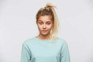 Closeup of unhappy upset young woman with blonde hair and ponytail wears blue sweatshirt feels displeased and looks to the camera isolated over white background photo