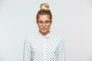 retrato de confidente atractivo joven mujer con bollo usa polca punto camisa y gafas siente grave y mira a el cámara aislado terminado blanco antecedentes foto